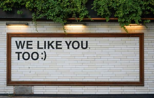 Outdoor sign on brick saying We Like You, TOO with a smiley emoticon